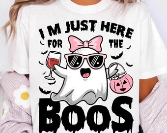 Op de afbeelding: Wit T-shirt met een Halloween-thema. Het ontwerp toont een cartoongeest met een zonnebril en een roze strik, die een wijnglas en een pompoenmand vasthoudt. De tekst luidt "I'M JUST HERE FOR THE BOOS."