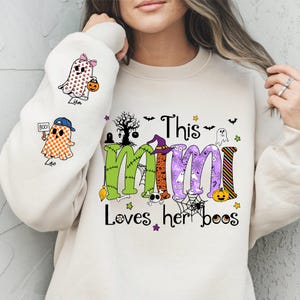 May include: Cream-colored sweatshirt with a Halloween-themed graphic. The design features the words "This Mimi Loves her boos" in green, orange, and purple, along with Halloween icons like ghosts, bats, and a witch's hat. The left sleeve has ghost illustrations.