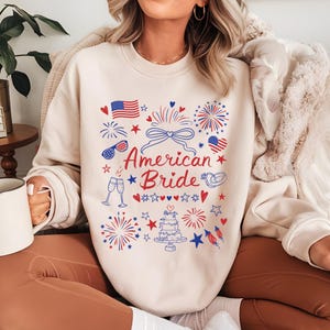 May include: A cream-colored sweatshirt with a patriotic design featuring the text "American Bride" in red and blue. The design includes fireworks, American flags, hearts, and a wedding cake. The sweatshirt is a casual, festive garment.