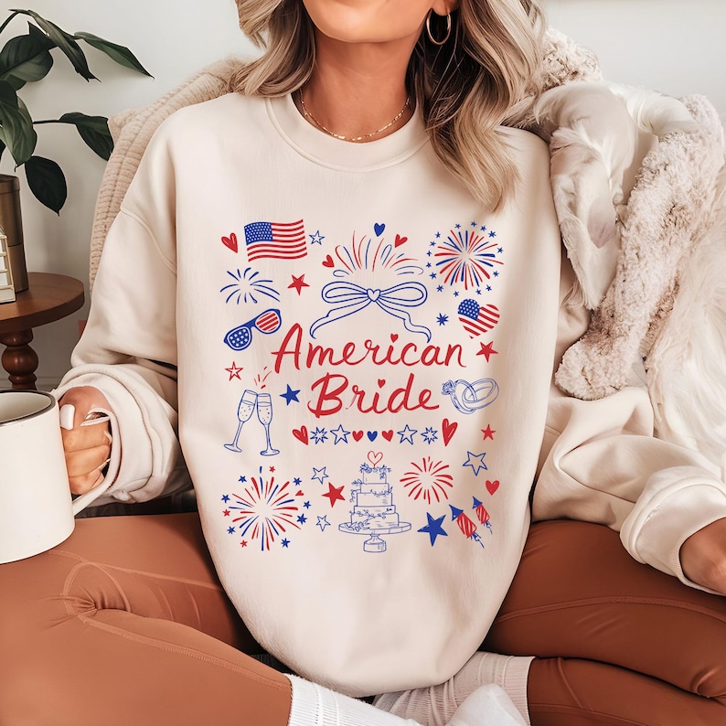 May include: A cream-colored sweatshirt with a patriotic design featuring the text "American Bride" in red and blue. The design includes fireworks, American flags, hearts, and a wedding cake. The sweatshirt is a casual, festive garment.