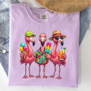 May include: A light purple Comfort Colors t-shirt featuring three cartoon flamingos. One flamingo wears a green cap and holds an ice cream cone, another wears a Hawaiian shirt and holds a popsicle, and the third wears a hat and sunglasses, holding a drink.