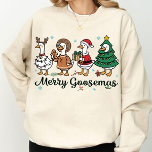 Peut inclure: Un sweat-shirt cr&egrave;me avec un motif festif. Quatre oies de dessin anim&eacute; sont habill&eacute;es pour les f&ecirc;tes. L'une porte des bois de renne, une autre tient un bonhomme en pain d'&eacute;pices, et une autre est habill&eacute;e en P&egrave;re No&euml;l. Le texte "Merry Goosmas" est imprim&eacute; en dessous.