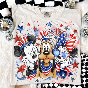 May include: White Comfort Colors t-shirt featuring Mickey Mouse, Pluto, and Minnie Mouse in patriotic attire. The design includes red, white, and blue stars, fireworks, and hats, perfect for Independence Day celebrations.