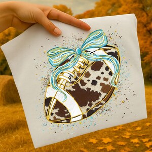 May include: A decorative football design with a cowhide pattern, white stripes, and gold accents. A light blue and gold bow adorns the top. The design is on a light beige background with gold and blue speckles.