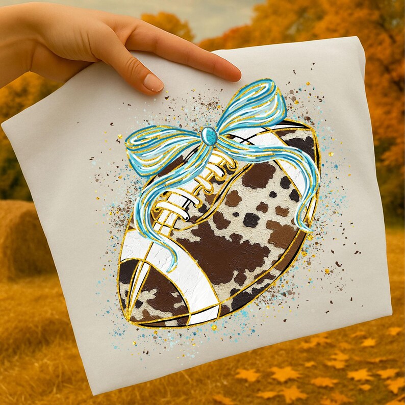 May include: A decorative football design with a cowhide pattern, white stripes, and gold accents. A light blue and gold bow adorns the top. The design is on a light beige background with gold and blue speckles.