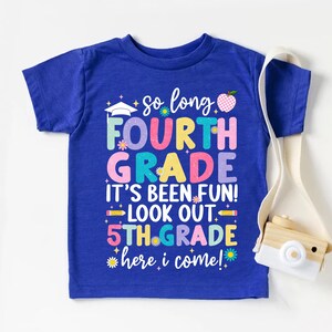 Puede incluir: Camiseta azul con el texto: "So long Fourth Grade, It's been fun! Look out 5th Grade, here I come!" Presenta un gorro de graduaci&oacute;n, una manzana y detalles florales. Un dise&ntilde;o colorido para el regreso a clases.