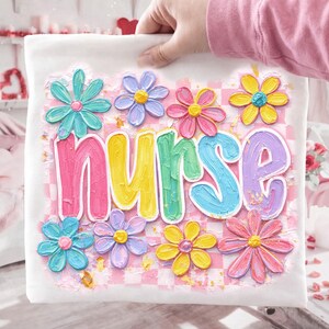 May include: White t-shirt with the word "NURSE" in colorful, textured letters, surrounded by pastel-colored flower designs. The background has a pink and white checkered pattern.