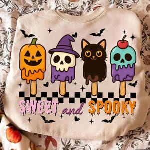 Op de afbeelding: Beige sweatshirt met een Halloween-thema. Het heeft vier personages in de vorm van ijslolly's: een pompoen, een schedel met een heksenhoed, een zwarte kat en een schedel met een kers. De woorden "SWEET and SPOOKY" staan eronder.