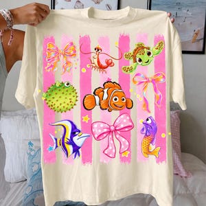 Peut inclure: Un t-shirt couleur crème arborant un imprimé graphique de coups de pinceau roses verticaux. Le motif comprend des illustrations d'un poisson-globe, d'un poisson-clown, d'une tortue de mer, d'une crevette, d'un poisson violet et trois nœuds décoratifs avec des étoiles jaunes.