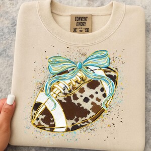 May include: Beige sweatshirt with a football design. The football has a cowhide pattern, white laces, and a blue and gold bow. The tag reads "CONVENT DIORY CO. AT TO IS."