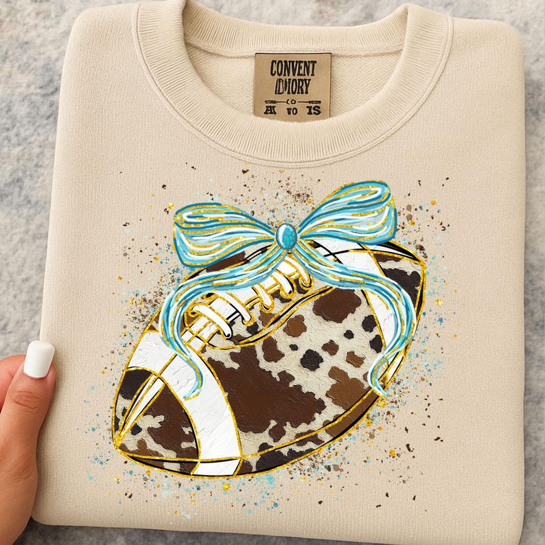 May include: Beige sweatshirt with a football design. The football has a cowhide pattern, white laces, and a blue and gold bow. The tag reads "CONVENT DIORY CO. AT TO IS."