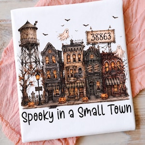 May include: A white t-shirt featuring a graphic illustration of a haunted town with a water tower, pumpkins, ghosts, and bats. A billboard displays the number 38863. The text Spooky in a Small Town is printed in black at the bottom of the design.