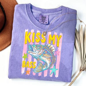 May include: A purple Comfort Colors t-shirt with the text "KISS MY BASS" in yellow letters. The graphic features a colorful fish with pink and green stars. A gold necklace is draped on the shirt.
