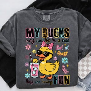 May include: Dark gray sweatshirt with the text "MY DUCKS may not be in a row But at least They are having FUN." Features a yellow duck wearing sunglasses and a pink bow, holding a drink with ice cubes.