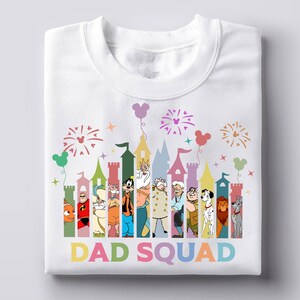 Pu&ograve; includere: Felpa bianca con una grafica colorata di un castello, personaggi Disney e fuochi d'artificio. Il testo "DAD SQUAD" &egrave; visualizzato a colori arcobaleno sotto il disegno del castello. La felpa &egrave; piegata.