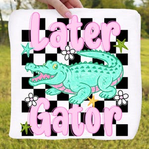 May include: White square towel with a cartoon alligator design. The alligator is teal with pink accents, set against a black and white checkered background. The words "Later Gator" are in pink script.