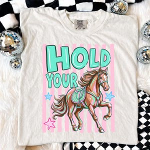 May include: Cream-colored t-shirt featuring a horse illustration with the text "HOLD YOUR". The horse is brown with a teal saddle and pink accents. Pink vertical stripes and blue stars surround the horse. The shirt has a Comfort Colors tag.