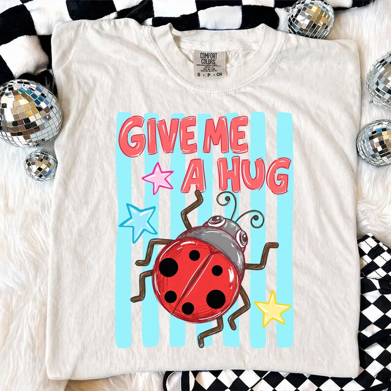 Pu&ograve; includere: T-shirt color crema con la scritta "GIVE ME A HUG" in lettere rosse. &Egrave; presente una coccinella dei cartoni animati con corpo rosso e pois neri, insieme ad accenti di stelle blu e gialle. La maglietta &egrave; del marchio Comfort Colors.