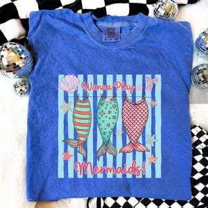May include: Blue Comfort Colors t-shirt with a graphic of three mermaid tails hanging on a clothesline. The text "Wanna Play Mermaids!" is above and below the tails. The tails are in shades of pink, green, and orange.