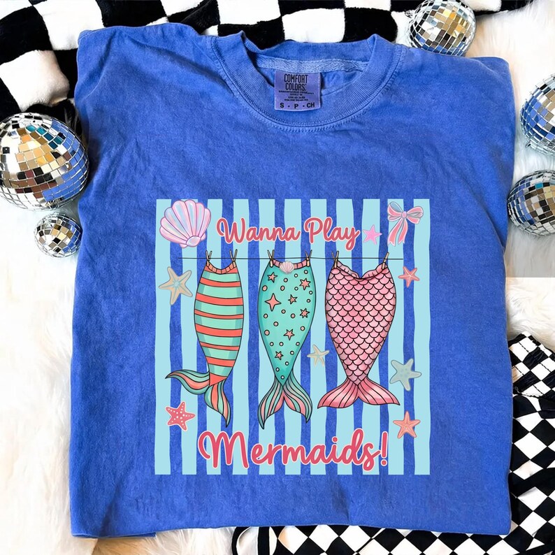 May include: Blue Comfort Colors t-shirt with a graphic of three mermaid tails hanging on a clothesline. The text "Wanna Play Mermaids!" is above and below the tails. The tails are in shades of pink, green, and orange.