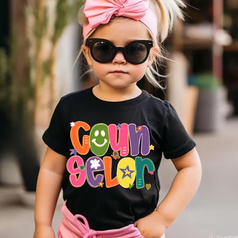 May include: A young child wearing a black t-shirt with the word "Counselor" in colorful, playful lettering. The child is also wearing sunglasses and a pink bow headband.
