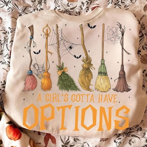May include: Cream-colored sweatshirt with a Halloween-themed design. The graphic features six different broom designs, bats, and spiderwebs. The text "A Girl's Gotta Have Options" is printed in orange block letters.