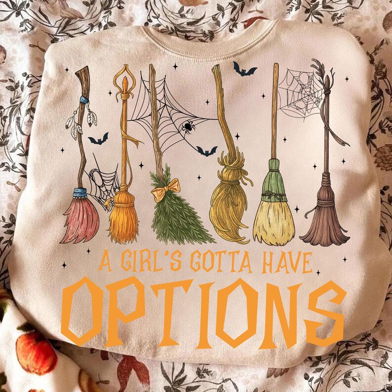 May include: Cream-colored sweatshirt with a Halloween-themed design. The graphic features six different broom designs, bats, and spiderwebs. The text "A Girl's Gotta Have Options" is printed in orange block letters.