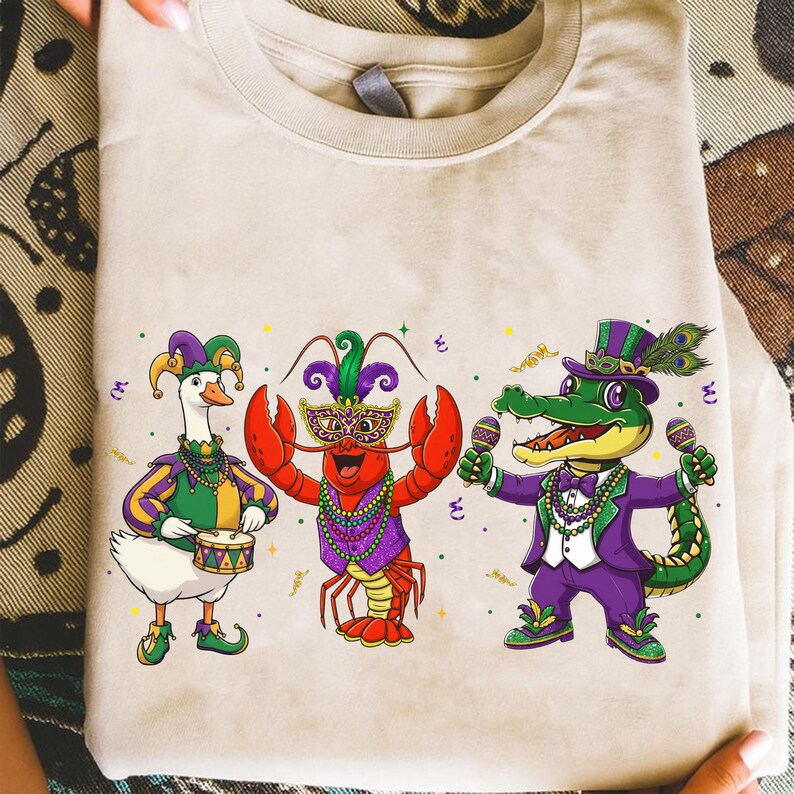 May include: Beige t-shirt featuring three cartoon characters in Mardi Gras attire. A goose plays a drum, a lobster wears a mask, and an alligator holds maracas. The characters are in green, purple, and gold.