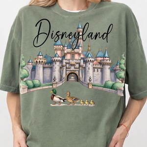 May include: Sage green t-shirt featuring a whimsical illustration of Disneyland's castle with ducks and ducklings. The text "Disneyland" is written in a cursive font above the castle. The shirt has short sleeves and a crew neck.