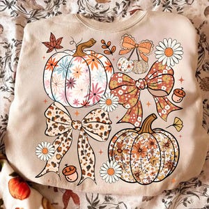 May include: Beige sweatshirt featuring a fall-themed design. The design includes two pumpkins with floral patterns, two bows, daisies, and acorns. The pumpkins have orange, white, and brown colors. The bows have a leopard print and floral patterns.