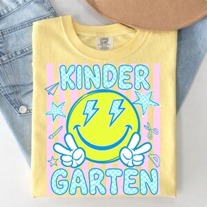 May include: Yellow t-shirt with a graphic design for kindergarten. The design features a smiley face with lightning bolts, peace signs, and the words "KINDER GARDEN" in a blue checkered font. Pink vertical stripes and school-themed illustrations complete the design.