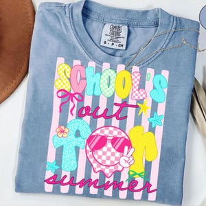 May include: A light blue Comfort Colors t-shirt with the words "Schools Out For Summer" in a colorful, playful design. The graphic includes a smiley face wearing sunglasses and various summer-themed elements.