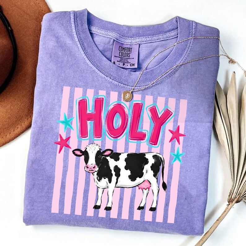 Puede incluir: Una camiseta de color lila con la palabra "HOLY" en rosa y azul, con una ilustraci&oacute;n de una vaca en blanco y negro. Estrellas rosas y azules rodean el texto. La camiseta tiene una etiqueta Comfort Colors.