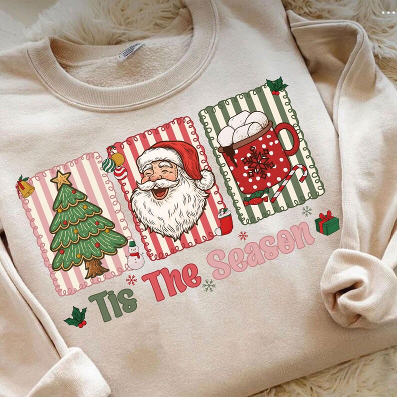 Puede incluir: Sudadera beige con un gr&aacute;fico festivo. El dise&ntilde;o incluye un &aacute;rbol de Navidad, Pap&aacute; Noel y una taza de chocolate caliente. Tambi&eacute;n se muestra el texto "Tis The Season". La sudadera tiene cuello redondo y mangas largas.