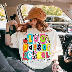 May include: Cream-colored sweatshirt with the text "100 Days of School" in colorful block letters. The design includes an apple, pencil, and a paper airplane. The person in the image is wearing a brown corduroy cap and sunglasses.