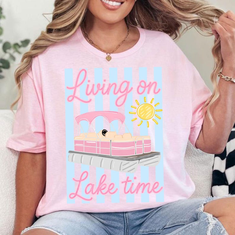 May include: Pink t-shirt with a graphic of a pink pontoon boat, a yellow sun, and the text "Living on Lake time" in pink script. The design is set against a light blue striped background.