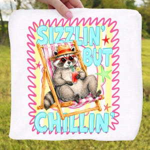 May include: White square pillow with a cartoon raccoon wearing sunglasses and a straw hat, relaxing in a beach chair. The pillow has the text "Sizzlin' But Chillin'" in blue and pink, with stars and a wavy border.