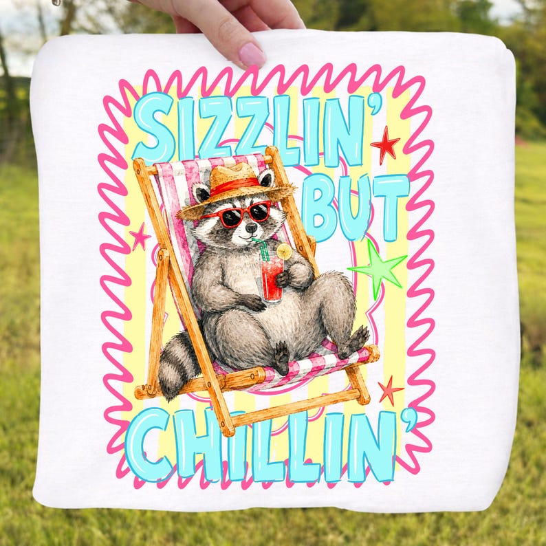 May include: White square pillow with a cartoon raccoon wearing sunglasses and a straw hat, relaxing in a beach chair. The pillow has the text "Sizzlin' But Chillin'" in blue and pink, with stars and a wavy border.