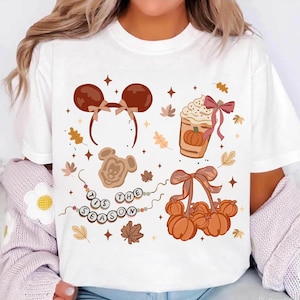 May include: White t-shirt featuring fall-themed graphics. The design includes a headband with mouse ears, a pumpkin spice latte, a Mickey Mouse-shaped cookie, a basket of pumpkins, and the phrase "IS THE SEASON".