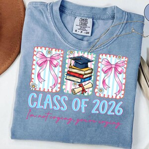 May include: A light blue t-shirt with a graduation theme. The shirt features three framed graphics with pink bows, books, a graduation cap, and a diploma. The text "CLASS OF 2026" is in light blue, with the phrase "I'm not crying, you're crying" below.