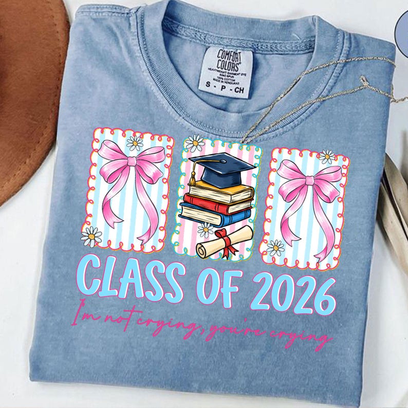 May include: A light blue t-shirt with a graduation theme. The shirt features three framed graphics with pink bows, books, a graduation cap, and a diploma. The text "CLASS OF 2026" is in light blue, with the phrase "I'm not crying, you're crying" below.