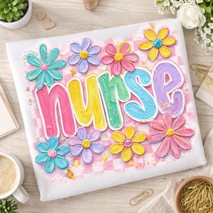 May include: White t-shirt with the word "NURSE" in colorful, textured lettering, surrounded by pastel-colored flowers. The background features a pink and white checkered pattern, creating a cheerful and artistic design.