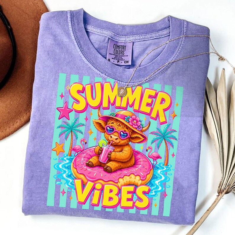 May include: A lilac-colored t-shirt with a graphic featuring a cartoon Highland cow wearing sunglasses and a hat, sitting on a donut float. The text "SUMMER VIBES" is displayed in bright yellow and pink. A gold necklace is draped on the shirt.