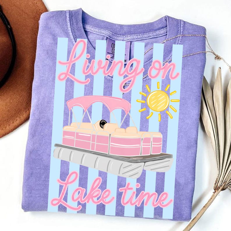 May include: A lilac-colored t-shirt with a graphic of a pink and white pontoon boat, a yellow sun, and the text "Living on Lake time" in pink script. The design is set against a backdrop of light blue vertical stripes.
