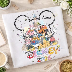 May include: White sweatshirt featuring a Disney design with Mickey Mouse, Minnie Mouse, Donald Duck, and other characters. The design includes the word "Disney" and the year "2026". A customizable "Name" is above the characters.