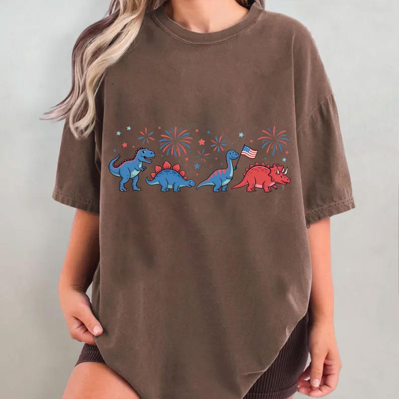 Puede incluir: Camiseta marr&oacute;n con una fila de dinosaurios de dibujos animados en azul y rojo, con fuegos artificiales y estrellas. Un dinosaurio sostiene una bandera americana. La camiseta tiene un corte holgado y mangas cortas, ideal para uso casual.