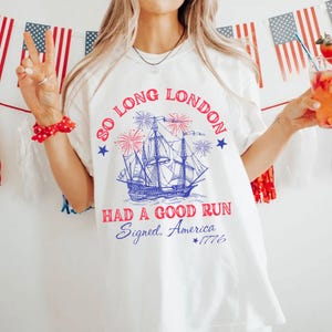 May include: White t-shirt with a patriotic design. The graphic features a blue ship, fireworks, and the text "SO LONG LONDON HAD A GOOD RUN Signed, America 1776" in red and blue. The shirt is a casual style.