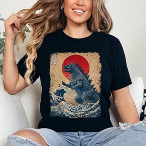 May include: Black t-shirt with a Godzilla-themed design. The graphic shows Godzilla in a traditional Japanese art style, with a red sun and ocean waves. The word "KAIJU" is printed vertically on the left side of the design.