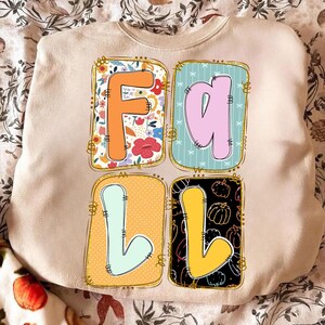 May include: Beige sweatshirt featuring the word "FALL" in a colourful, decorative design. The "F" is orange with a floral pattern, the "A" is pink with blue stripes, the first "L" is light blue on yellow, and the second "L" is yellow with pumpkins.