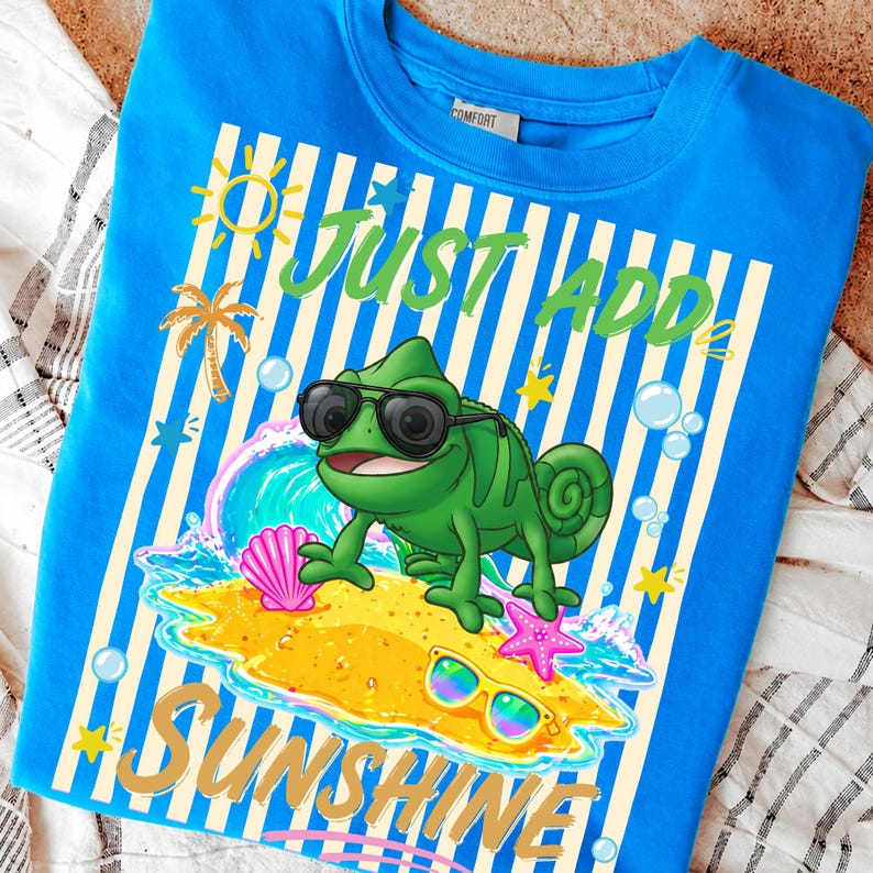 May include: A blue t-shirt with a graphic featuring a green chameleon wearing sunglasses on a beach with the text "Just Add Sunshine". The design includes a sun, palm tree, and other beach elements.